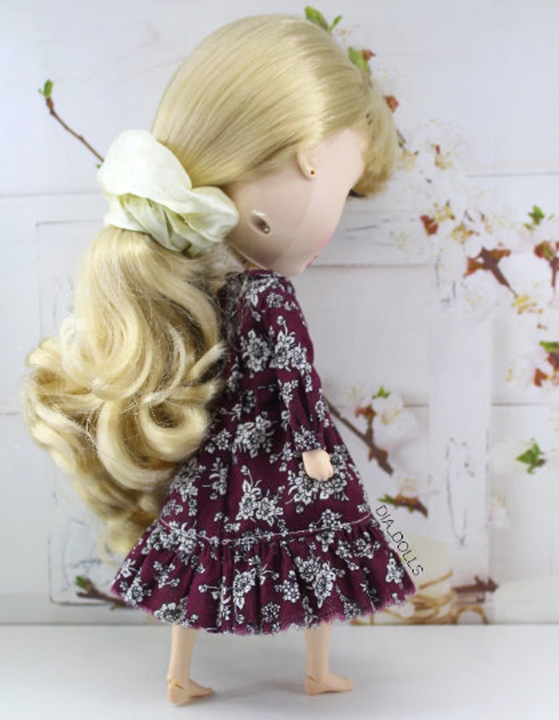 May include: A doll with long blonde hair styled in a ponytail with a white scrunchie. The doll is wearing a burgundy dress with a white floral pattern and a ruffled hem. The doll is standing on a white surface.