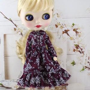 May include: A doll wearing a burgundy dress with a white floral pattern. The dress has a ruffled hem and long sleeves. The doll has blonde hair and blue eyes.