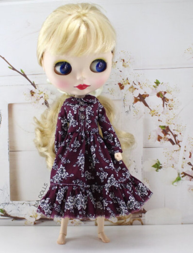 May include: A doll wearing a burgundy dress with a white floral pattern. The dress has a ruffled hem and long sleeves. The doll has blonde hair and blue eyes.