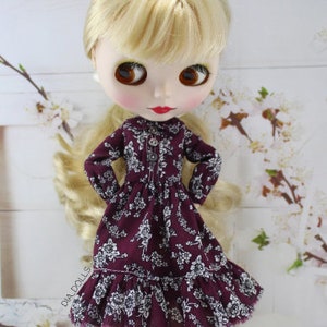 May include: A doll wearing a burgundy dress with white floral print and a ruffled hem. The doll has blonde hair and brown eyes.