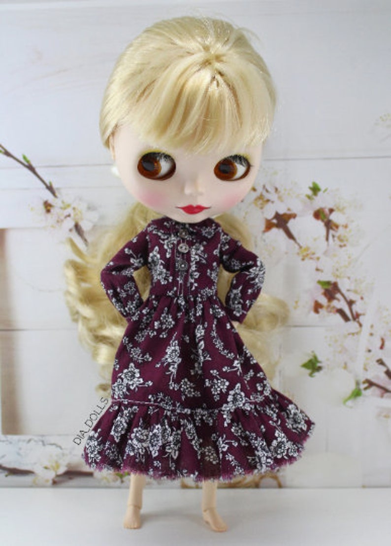 May include: A doll wearing a burgundy dress with white floral print and a ruffled hem. The doll has blonde hair and brown eyes.