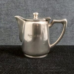 Puede incluir: Una pequeña cremera plateada con tapa y asa. La cremera tiene un pico para verter y el texto "Kursaal Bern" es visible en la parte delantera. La cremera es probablemente de metal y se encuentra sobre una superficie oscura.
