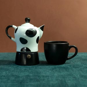 Puede incluir: Una cafetera italiana blanca y negra con lunares, con un asa y un pico negros, junto a una taza de café negra maciza. La cafetera tiene una base negra y un pequeño botón dorado.