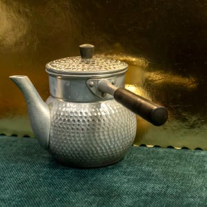 Peut inclure: Une théière en argent avec une finition martelée et une poignée noire. La théière a un couvercle avec un petit bouton sur le dessus.