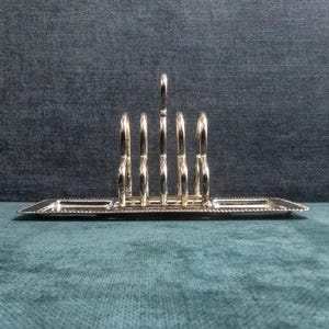 May include: A silver-colored toast rack with six arched slots and a rectangular base. The rack has a central handle and a beaded edge, suitable for serving toast at the breakfast table.