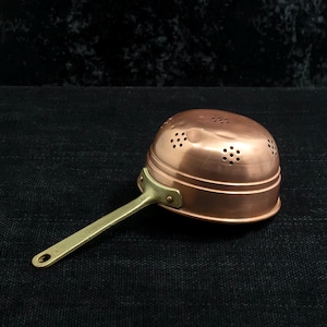 May include: A copper colander with a rounded bowl shape featuring multiple small circular drainage holes across the top and sides. The colander includes a long, straight brass handle attached to the side with two visible metal rivets.