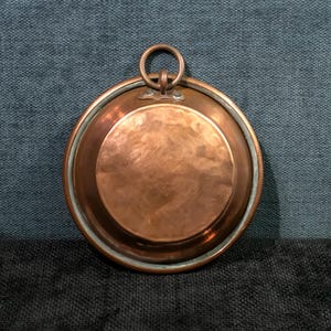 Pode incluir: Uma panela redonda rasa, cor de cobre, com uma borda elevada e uma pequena alça circular. A panela tem uma superfície polida e uma pátina escura e envelhecida. A alça é presa à panela com um suporte decorativo.