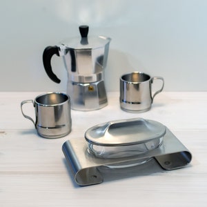 May include: A silver coffee pot with two silver mugs and a silver lidded dish on a white wooden surface.
