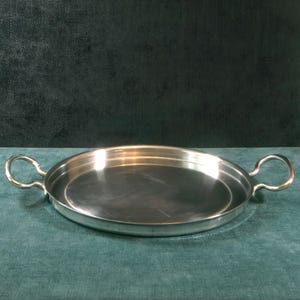May include: A shiny, silver-colored serving tray with two loop handles. The tray is oval-shaped and has a slightly raised rim. The surface reflects light, suggesting a polished finish. The tray is likely made of metal and is suitable for serving food or displaying items.