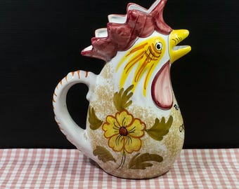 Puede incluir: Una jarra de cerámica con forma de gallo con base blanca, cresta roja y pico amarillo. El cuerpo presenta una flor amarilla pintada a mano con hojas verdes y una textura marrón moteada. Un asa curva está unida al costado de la jarra.