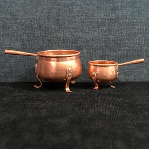 May include: Two copper pots with handles and curved legs. The larger pot has a diameter of approximately 6 inches, while the smaller pot is about 3 inches in diameter. Both pots have a shiny, reflective surface and are designed for cooking.