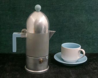 May include: A silver stovetop espresso maker with a light blue handle and knob, next to a white cup and saucer with a light blue interior and rim. The coffee maker and cup are on a dark surface against a green backdrop.