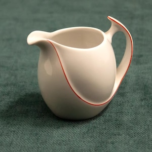 Può includere: Una lattiera in ceramica bianca con un manico curvo e un beccuccio. La lattiera ha un bordo rosso che delinea il manico e il corpo. La lattiera è su una superficie verde.