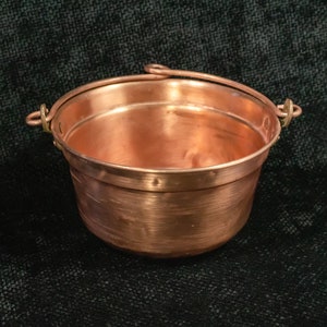 May include: A copper pot with a handle, the pot has a hammered finish and is sitting on a dark background.