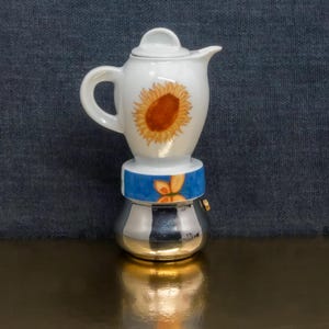 May include: A small espresso maker with a white ceramic top decorated with a sunflower. The base is silver with a gold-coloured bottom, and a blue band with a butterfly design separates the top and bottom. A charming kitchen item.