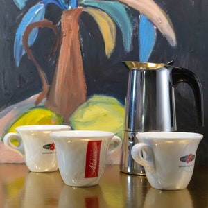 Könnte beinhalten: Drei weiße Espressotassen aus Keramik mit den Worten "Essai Espresso" und "Miasini" darauf gedruckt. Im Hintergrund steht eine silberne Moka-Kanne.