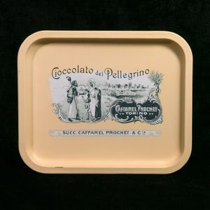 Può includere: Un vassoio in metallo vintage con sfondo crema e un'illustrazione in bianco e nero di persone in un paesaggio. Il testo "Cioccolato del Pellegrino" e "Caffarel Prochet Torino" è stampato sul vassoio.