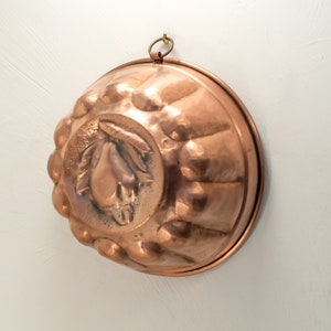 Op de afbeelding: Een ronde, vintage koperen vorm voor het maken van desserts. De vorm heeft een reliëfdesign van een peer met bladeren in het midden. De vorm heeft een ophanglus aan de bovenkant.