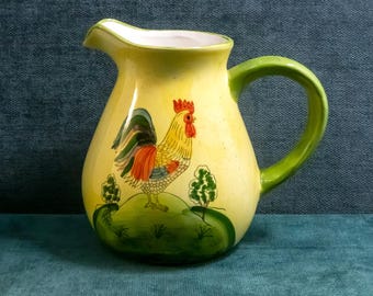 Puede incluir: Una jarra de cerámica amarilla con un gallo pintado a mano sobre una colina verde con dos árboles pequeños. La jarra tiene un asa verde curva, un pico ancho y un interior blanco, con un acabado moteado sobre la superficie amarilla.