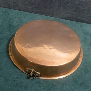 Op de afbeelding: Een ronde, koperkleurige pan met een licht verhoogde rand en een klein handvat met een ring. De pan heeft een gepolijst oppervlak en staat tegen een turquoise achtergrond.