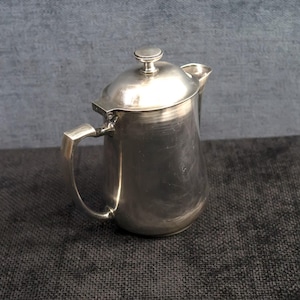 Puede incluir: Una jarra de plata con tapa y asa. La jarra está sobre una superficie gris oscura.