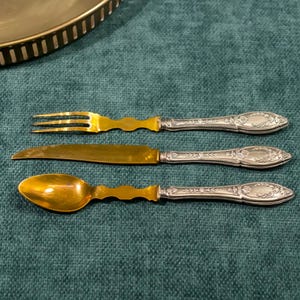 Può includere: Un set di tre posate in argento placcato oro, composto da una forchetta, un coltello e un cucchiaio. I manici sono in argento e presentano un motivo floreale decorato.