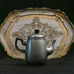 May include: A small, silver-colored metal teapot with a black handle and lid knob. The teapot is set against a decorative, ornate tray with a white and gold design. The background is a dark, textured surface.
