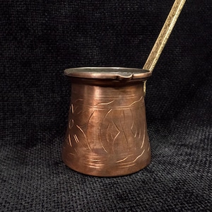 May include: A copper coffee pot with an ornate design and a brass handle. The pot is on a black background.
