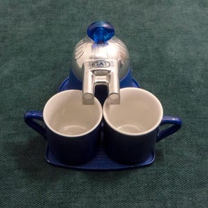 Op de afbeelding: Een blauwe espressomaker met twee bijpassende blauwe kopjes. De espressomaker is zilver en blauw met het woord "GAT" op de voorkant. De kopjes staan op een blauw dienblad.