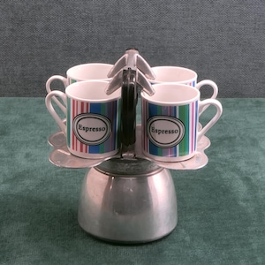 May include: A set of four white ceramic espresso cups with vertical stripes in shades of blue, green, red, and purple. Each cup features a black oval emblem with the word Espresso printed in white. The cups are arranged on a metal stand.