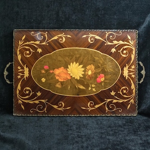 Peut inclure: Un plateau de service rectangulaire avec une finition en grain de bois foncé, présentant une incrustation ovale centrale d'un bouquet floral dans des tons orange, jaune et rose. Le plateau est bordé de motifs en filigrane dorés et possède deux poignées en métal ornées.