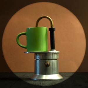 Op de afbeelding: Een groene keramische mok staat op een zilveren espressomaker. De espressomaker heeft een zwarte handgreep en een goudkleurige tuit. De achtergrond is warmbruin met een spotlight-effect.