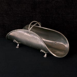 May include: A silver sugar scuttle with a curved body and a twisted rope-like handle. The scuttle has two small, decorative feet and a rope-like trim around the rim. The background is black.