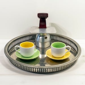 Puede incluir: Una bandeja de plata con una cafetera de plata y roja, dos tazas de café blancas con platillos amarillos y verdes.