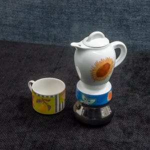 May include: A white ceramic espresso maker with a sunflower design, a blue band with floral accents, and a silver base. A small cup with a colorful fruit and stripe pattern sits beside it.