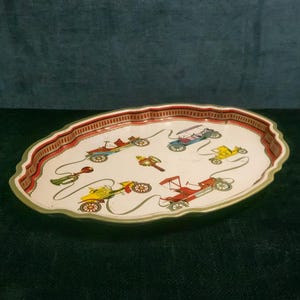 May include: Oval-shaped vintage metal tray with a cream-colored surface. The tray features a repeating pattern of colorful antique cars in red, yellow, blue, and green. The tray's edge has a red and tan striped border with a green trim.