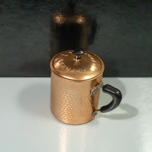 Könnte beinhalten: Eine Kaffeekanne aus Kupfer mit schwarzem Griff und Deckel. Der Deckel hat das Wort "Express" geprägt.