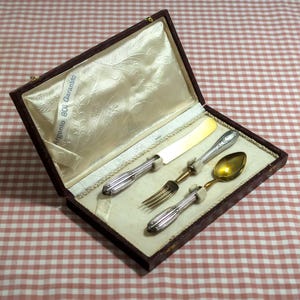 May include: A vintage silverware set in a brown box with a cream-colored satin lining. The set includes a knife, fork, spoon, and a smaller utensil. The handles are silver, and the spoon has a gold-colored bowl. The box has the text "Argento 800 Garantito".