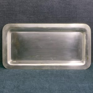 Peut inclure: Un plateau rectangulaire en métal argenté doté d'une base lisse et plate avec des bords larges et arrondis. La surface présente une finition polie et réfléchissante avec de légères variations de ton, offrant un design épuré et fonctionnel.