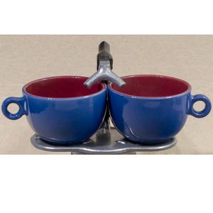 May include: A blue ceramic double cup set with red interiors and loop handles. The cups are connected by a metal fixture with a spout. The set is designed to sit on a metal base.