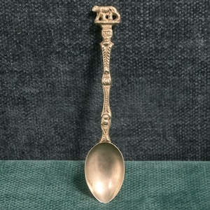 May include: A silver-colored souvenir spoon with a detailed handle and a bear figure at the top. The spoon has an ornate design and a rounded bowl. The spoon is displayed against a dark teal background.