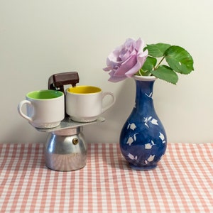 Könnte beinhalten: Eine weiße und grüne Kaffeetasse, eine weiße und gelbe Kaffeetasse und eine silberne Kaffeekanne auf einer weißen und rosa karierten Tischdecke. Eine blaue Vase mit weißen Blumendesigns hält eine einzelne lila Rose.