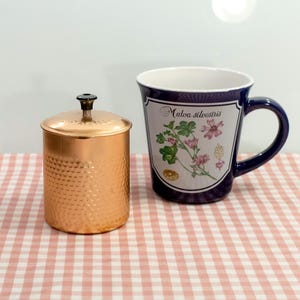 May include: A copper container with a lid and a blue ceramic mug with a floral design and the text "Malva silvestris".