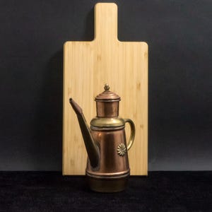 Op de afbeelding: Een koperen en messing olie schenker met een lange tuit en een klein handvat, geplaatst voor een lichtgekleurde houten snijplank. De olie schenker heeft een decoratief bloemmotief aan de zijkant. De achtergrond is zwart.