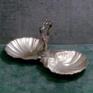 Könnte beinhalten: Ein zweiteiliges Serviertablett in Silberoptik in Form von Muscheln. Das Tablett hat einen dekorativen Griff in der Mitte. Das Tablett besteht aus glänzendem, reflektierendem Metall.