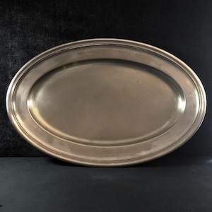 May include: An oval-shaped silver-toned metal serving platter with a smooth, polished surface and a raised, rounded rim. The platter features a simple, clean design with a reflective finish that catches the light, creating subtle highlights along the edge.
