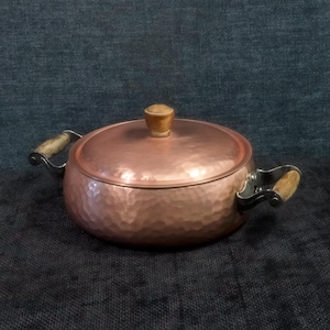 May include: A hammered copper pot with a wooden knob lid and wooden handles. The pot has a rounded shape and a textured surface.