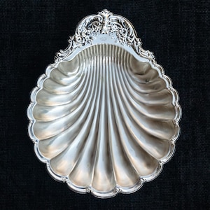 Puede incluir: Un plato de metal plateado con forma de concha de vieira. El interior presenta profundas ranuras radiantes, mientras que el borde superior está decorado con un patrón de volutas en relieve. La superficie tiene un acabado pulido y reflectante.