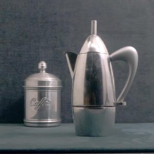 Pode incluir: Uma cafeteira italiana prateada e um recipiente a condizer. A cafeteira tem um topo arredondado e uma pega curva. O recipiente tem uma tampa e a palavra "Caffe" em relevo na frente.