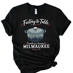 May include: A black short-sleeve t-shirt featuring a graphic of a light blue Dutch oven pot with a city skyline inside. The text reads Factory to Table, Something's brewing in MILWAUKEE, accented with small red hearts and decorative lines.